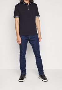 Polo shirt marine avec fermeture éclair, manches courtes, présentant des accents blancs contrastants sur le col et les ourlets des manches, associé à un jean bleu foncé et des baskets noires.