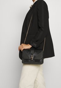 Czarna torebka z teksturowanym wzorem, łańcuszkowym paskiem i metalowym zapięciem. Prostokątny kształt z kontrastowymi materiałami na klapie.