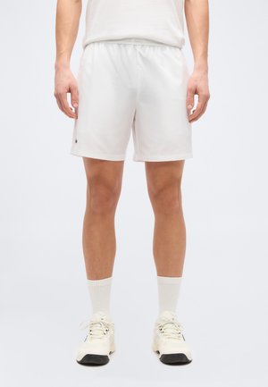 Fehér atlétikai rövidnadrágot, fehér bokazoknit és fehér tornacipőt viselő férfi, egyenes tartással, egy egyszínű világos háttér előtt áll.