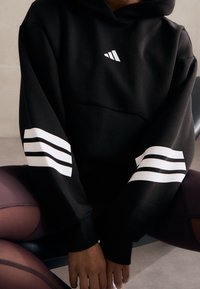 Czarna bluza z kapturem z białym logo Adidas i trzema białymi paskami na każdym rękawie. Wykonana z miękkiej, teksturowanej tkaniny.