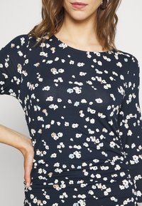 Haut floral marine en tissu doux ; présente de petites fleurs blanches, bleues et brunes ; coupe décontractée et col rond avec un détail de ruché sur les côtés.