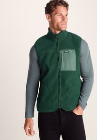 Man wearing a dark green fleece vest with front zipper and chest pocket over a gray long-sleeve shirt, standing against a plain background.