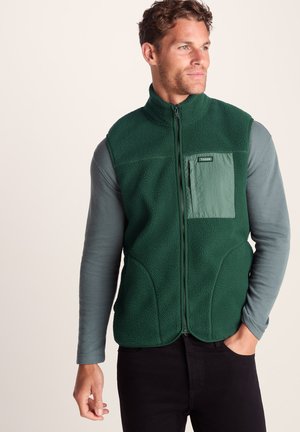 Man wearing a dark green fleece vest with front zipper and chest pocket over a gray long-sleeve shirt, standing against a plain background.