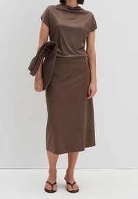 Haut à manches courtes marron et jupe longue marron, tous deux en tissu lisse. Le mannequin tient un châle marron assorti. Les sandales présentent un design simple.