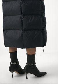 Černý péřový kabát s prošívaným vzorem a bočním zipem; černé kotníkové boty s špičatou špičkou, řetízkovým detailem a vysokým jehlovým podpatkem.