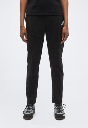 Persoană purtând pantaloni cargo negri cu buzunare cu fermoar și pantofi de drumeție gri, stând în față unui fundal alb simplu.