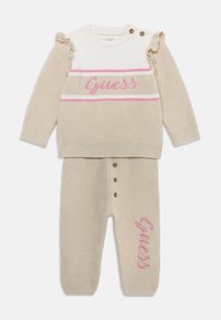Beige gestricktes zweiteiliges Outfit mit einem Oberteil mit pinkem Logo und Rüschärmeln sowie passenden Hosen mit Knopfdetails und Logoprinz. 