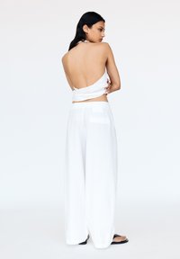 Weißes Halterneck-Top mit offenem Rücken, kombiniert mit weitem weißen Hosen. Glattes Material, leichtes Textil, minimalistisches Design. Schwarze Sandalen.