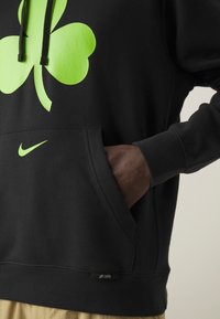 Sweat à capuche noir avec un trèfle néon vert et un logo Nike, main dans la poche avant, et une étiquette "City Edition" à l'ourlet.