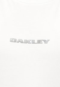 Λευκό υφασμάτινο φόντο με κεντρικό ανάγλυφο ασημένιο κείμενο που γράφει "OAKLEY" με έντονα κεφαλαία γράμματα.