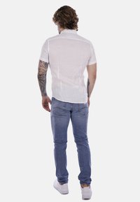 Camicetta bianca di lino a maniche corte, vista dal retro, abbinata a jeans azzurri e sneakers bianche. Presenta una texture morbida, un design minimalista e una vestibilità casual.