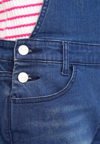 Dark blue denim overalls feature silver hardware, two buttons, and a front pocket with a curved seam. The texture appears smooth with slight fading.