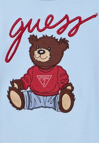 Ruskea nalle, jolla on punainen neulepaita valkoisella "Guess"-logolla ja siniset housut, istuu suuren punaisen kursiivitekstillä "guess" varustetun vaaleansinisen taustan alla.