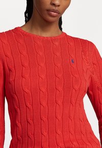 Frau trägt einen roten Strickpullover mit Zopfmuster und einem kleinen blau bestickten Logo auf der Brust, vor einem schlichten weißen Hintergrund.