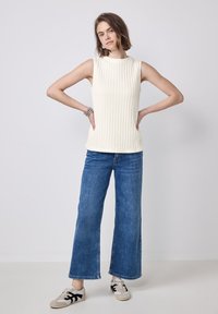 Ribbede, ærmeløse, elfenbensfarvede toppe med høj hals, kombineret med brede denimjeans og afslappede sneakers. Et armbånd på håndleddet tilføjer detaljer.