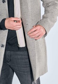 Hellgrauer Fischgrätmantel mit Reißverschluss und dunkelgrauem Pullover darunter, kombiniert mit dunkelgrauen Jeans. Nahaufnahme der Hände in der Nähe des Reißverschlusses.