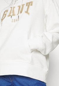 Sudadera blanca de tejido suave con la palabra bordada "GANT" y "1949" en dorado. Incluye un bolsillo frontal y puños de canalé.