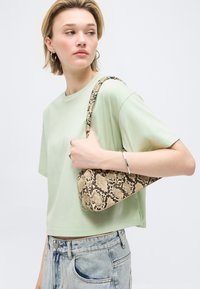 Νεαρή γυναίκα που φοράει ανοιχτό πράσινο κοντό μπλουζάκι και ανοιχτό μπλε τζιν, κρατώντας μια τσάντα ώμου με σχέδιο δέρματος φιδιού στον δεξιό της ώμο.