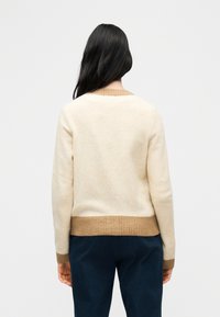 Γυναίκα με μακριά μαύρα μαλλιά, που φοράει μπεζ πουλόβερ με καφέ μανσέτες και τελείωμα, στέκεται απέναντι σε λευκό φόντο.