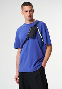 Schwarze Umhängetasche mit Logo, getragen an einem schwarzen Riemen über einem blauen T-Shirt mit kurzen Ärmeln. Einfaches Design mit glatter Textur.