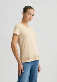 Korte mouw beige T-shirt van zachte stof, met een gemêleerde zoom en een subtiel patroon. Gecombineerd met lichtblauwe jeans.