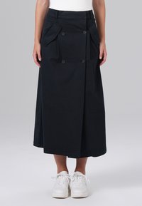 Jupe midi noire à design portefeuille, dotée de deux grandes poches avant, fermeture à double bouton et texture en coton lisse. Associée à des baskets blanches.
