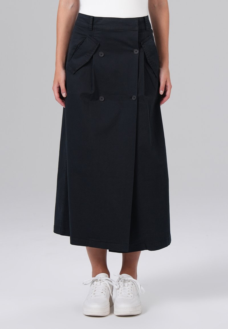Jupe midi noire à design portefeuille, dotée de deux grandes poches avant, fermeture à double bouton et texture en coton lisse. Associée à des baskets blanches.