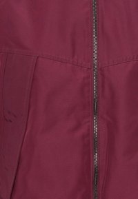 Veste bordeaux avec fermeture éclair, présentant une texture douce et légère ainsi que des détails de coutures. Le tissu a un éclat subtil.