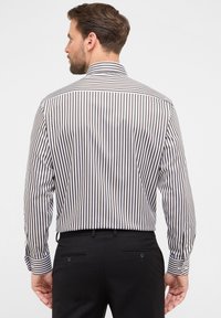 Man met kort bruin haar en baard, gekleed in een bruin-wit verticaal gestreept overhemd met lange mouwen en zwarte broek, gezien van achteren.