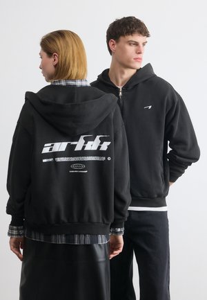 Dva mlada odrasla oseba, oblečena v črne kapuce; eden gleda naprej z minimalnim logotipom, drugi pa gleda nazaj, kjer je na hrbtu viden velik bel napis.