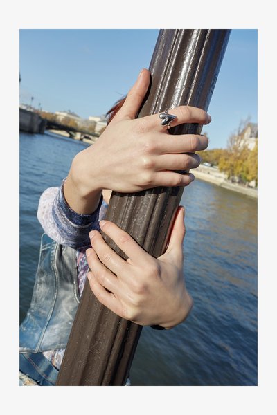 Silberner Ring mit Herzdesign an einer Hand, die einen braunen, strukturierten Pfahl am Wassergriff hält. Der Hintergrund zeigt Wasser und entfernt stehende Gebäude.
