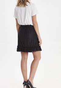 Czarna, fakturowana spódnica z falbanką i elastycznym pasem, noszona na białej koszulce. Widok z tyłu pokazujący kształt i dopasowanie.