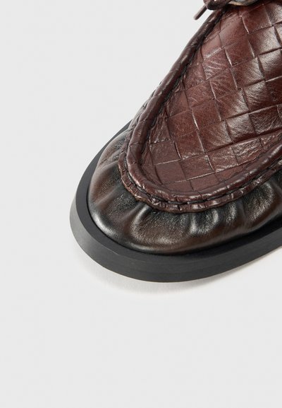 Scarpa in pelle marrone con una texture intrecciata sulla parte superiore, punta arrotondata e suola in gomma nera. Presenta dettagli arricciati attorno al bordo.