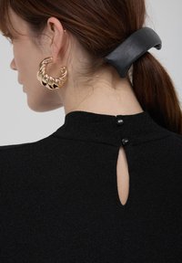 Frau mit braunen Haaren, die mit einer großen schwarzen Haarklammer zusammengebunden sind, trägt ein strukturiertes schwarzes Oberteil mit Knopfverschluss im Rücken und große goldene Creolen.