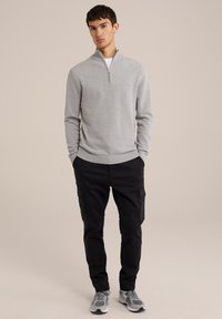 Hellgrauer Zip-Pullover mit gerippten Bündchen und Saum, kombiniert mit schwarzen Cargo-Hosen mit seitlichen Taschen und grauen Sneakers.