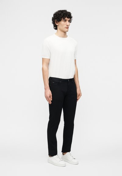 Άνδρας με σγουρά μαύρα μαλλιά, φορώντας ένα απλό λευκό κοντομάνικο T-shirt, μαύρο παντελόνι στενής εφαρμογής και λευκά αθλητικά παπούτσια, στέκεται μπροστά από ένα λευκό φόντο.