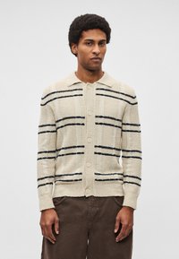 Bēša adīta kardigans ar melnām horizontālām svītrām, ar pogām aiztaisāms priekšpusē, apkakli un teksturētu audumu. Akcenti ietver apaļas pogas un kabatas.