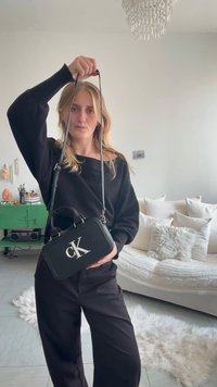 Γυναίκα που φορά μαύρα κρατά μια μαύρη τσάντα Calvin Klein από το λουρί της σε ένα μοντέρνο καθιστικό με λευκό καναπέ και πράσινη ντουλάπα.