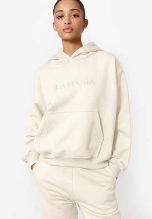 Młoda kobieta w kremowej bluzie z kapturem z napisem „LA MANIA” oraz pasujących kremowych spodniach dresowych, stojąca z rękami w kieszeniach na białym tle.