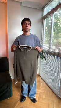 Homme debout à l'intérieur tenant un polo marron à manches longues avec un bord blanc près d'une fenêtre avec des stores.