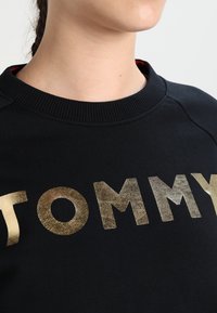 Felpa nera con collo a girocollo a coste e audace scritta in oro metallico che spellala "TOMMY" sul petto. Textura liscia.