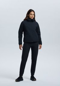 Zwarte geïsoleerde jas met textuurstof en een rits, gecombineerd met aansluitende zwarte broek en zwarte sportieve schoenen.