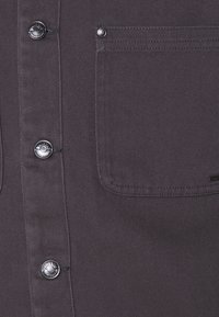 Koksgrå skjorte med knapper i metal med trykknapper og en brystlomme med diskrete syninger. Stoffet har en diagonal tekstur.