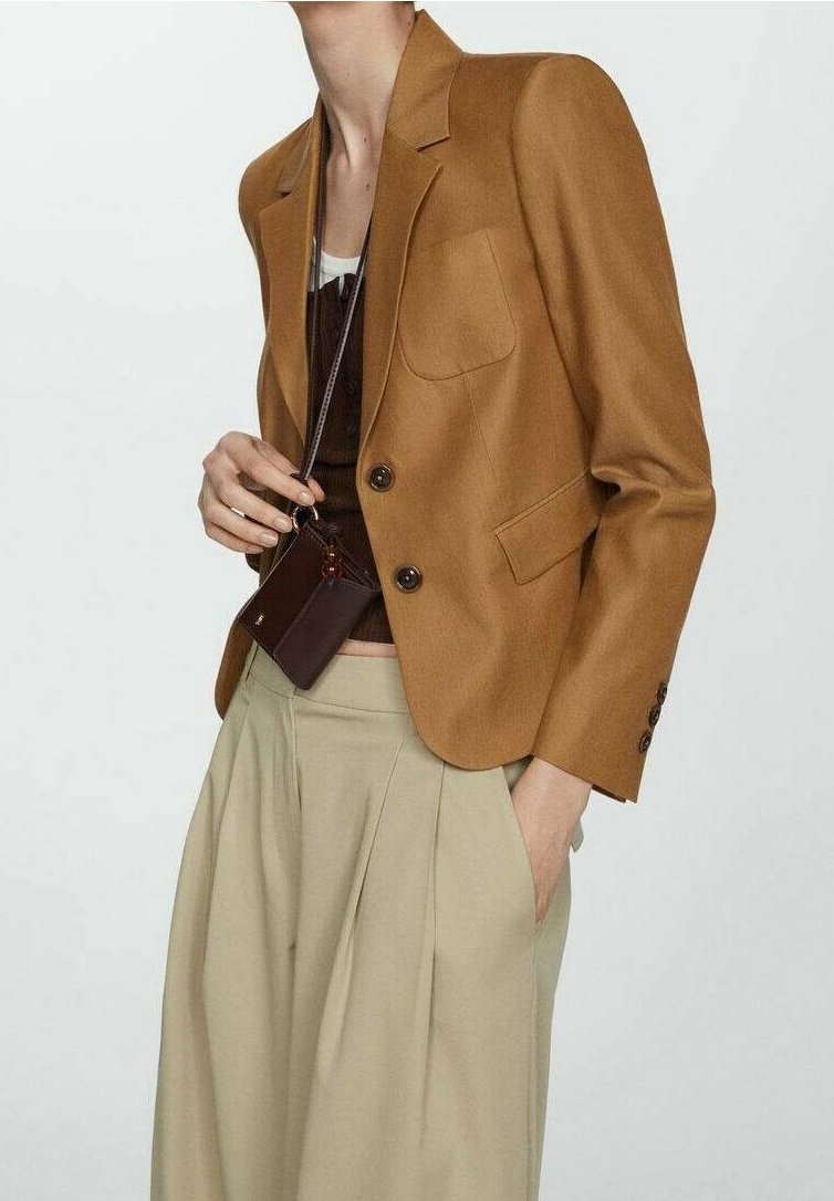 Personne portant un blazer court marron, un haut marron foncé, un pantalon beige taille haute à jambes larges, tenant un petit sac bandoulière marron foncé.