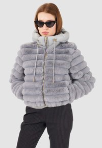 Giacca puffer grigia con texture a strisce morbide, dotata di cappuccio, zipper dorata e lacci. Indossata con pantaloni neri a strisce sartoriali.