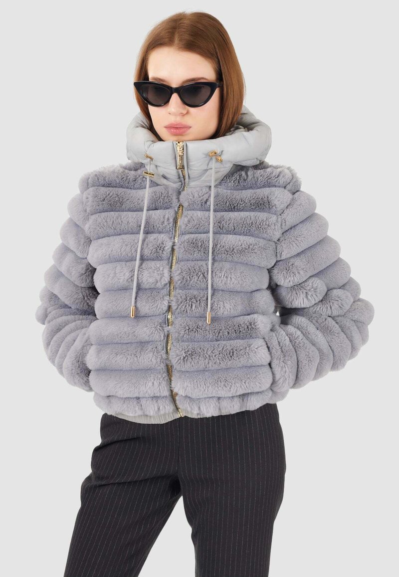 Giacca puffer grigia con texture a strisce morbide, dotata di cappuccio, zipper dorata e lacci. Indossata con pantaloni neri a strisce sartoriali.
