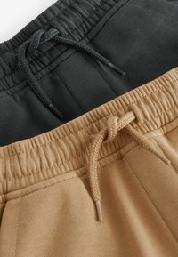 Dos pares de pantalones de chándal: uno en negro con una cinturilla texturizada y cordón, el otro en color tostado con una cinturilla lisa y costuras detalladas.