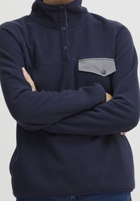 Sudadera de forro polar azul marino con cuello alto, tres botones de presión negros y un bolsillo en el pecho gris con detalle de botón. Textura suave.