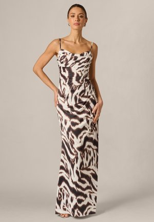 Frau steht und trägt ein ärmelloses, bodenlanges Kleid mit schwarz-weißem Tiermuster vor einfarbigem Hintergrund.