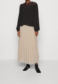 Blusa nera a maniche lunghe con yoke in pizzo, abbinata a una gonna plissettata metallic oro e scarpe sportive nere con suola bianca.